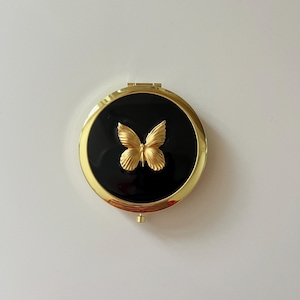 May include: A round, gold-toned metal compact mirror featuring a glossy black circular inlay on the lid. A three-dimensional, textured gold butterfly emblem is centered on the black surface. The compact includes a small latch for opening.