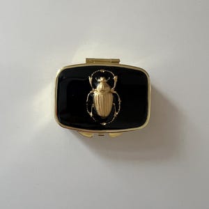 May include: A rectangular, hinged trinket box with a black enamel finish. The box features a gold-colored scarab beetle design on the lid. The box has gold-colored trim and a gold-colored hinge.