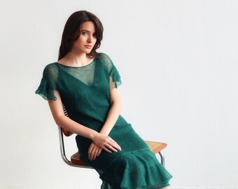 May include: A dark emerald green dress with short sleeves and a ruffled hem. The dress is paired with leopard print heels. The model is seated on a chair with a woven seat and metal frame. The dress appears to be made of a sheer, textured fabric.