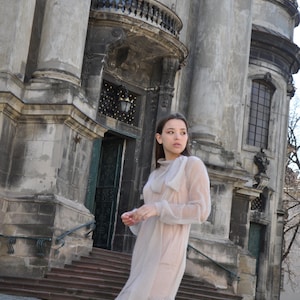 Può includere: Una donna che indossa un abito di tulle beige chiaro con un fiocco al collo, in piedi di fronte a un grande edificio decorato con una facciata in pietra.