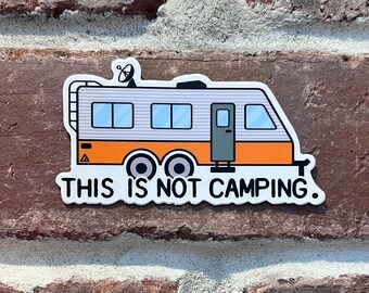 May include: A white sticker with a cartoon illustration of a grey and orange camper with a satellite dish on top. The text "THIS IS NOT CAMPING." is printed below the camper.
