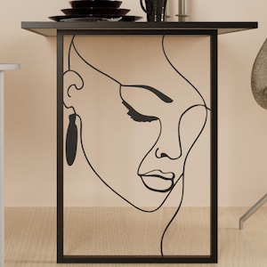 May include: Black metal side table with a minimalist design. The table features a black frame with a graphic outline of a woman's face. The table has a black top with black plates and a mug.