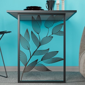 Könnte beinhalten: Ein moderner Beistelltisch mit schwarzem Metallrahmen und Blattdesign auf türkisfarbenem Hintergrund. Der Tisch hat eine dunkle Oberseite mit einer schwarzen Tasse, Tellern und Kerzen. Der Tisch ist etwa 61 cm hoch.