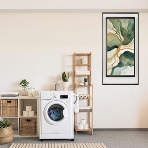 Peut inclure: Une buanderie avec une machine à laver blanche, des étagères en bois et une œuvre d'art abstraite encadrée aux tons verts et dorés. La pièce comprend également des plantes, des paniers et un tapis rayé, créant un espace propre et organisé.