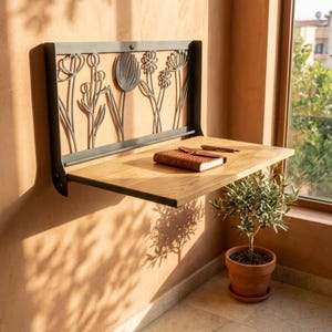 May include: A wall-mounted folding desk featuring a natural wood surface and a black metal frame with a decorative cutout design of various wildflowers. A brown leather-bound notebook and a pen rest on the flat wooden tabletop.
