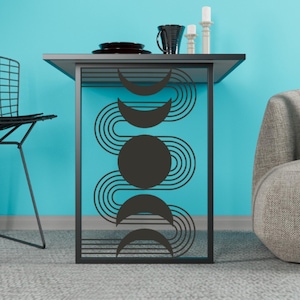 Op de afbeelding: Een zwarte metalen bijzettafel met een geometrisch ontwerp op een turquoise achtergrond. Het ontwerp toont gebogen lijnen en maanfasen. De tafel heeft een platte bovenkant met een zwarte mok, borden en kaarsen. De tafel is ongeveer 60 cm hoog.
