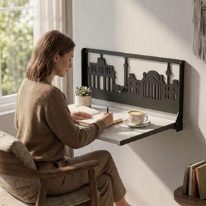 Op de afbeelding: Een aan de muur gemonteerd opklapbureau met een zwart metalen frame met een lasergesneden silhouet van de skyline van Berlijn, inclusief de Brandenburger Tor en het woord BERLIN, gecombineerd met een wit tafelblad om aan te werken.