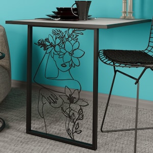 Könnte beinhalten: Kleiner, quadratischer Tisch mit schwarzem Metallrahmen und hellgrauer Platte. Die Seite des Tisches zeigt eine schwarze Strichzeichnung einer Frau mit Blumen. Ein schwarzer Stuhl und Geschirr sind sichtbar.