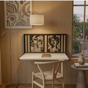 May include: A wall-mounted white desk featuring a black metal back panel with two distinct abstract face illustrations. The desk is positioned below a floor lamp with a beige shade and is accompanied by a light-colored wooden chair.