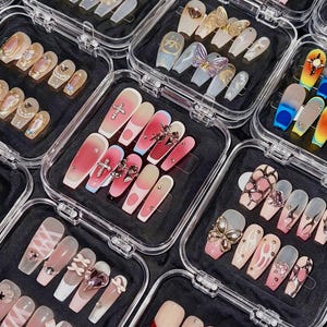 May include: Assortment of artificial nails in clear plastic cases. Designs include ombre, glitter, and 3D embellishments like crosses, butterflies, and gems. Colors range from pinks and reds to clear and iridescent.