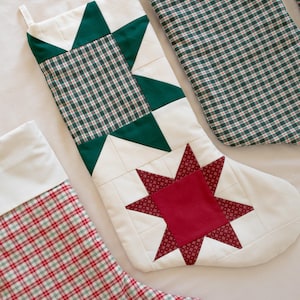 May include: Three quilted Christmas stockings featuring patchwork designs. One stocking has a green and white plaid pattern with a green star, another has a red and white plaid pattern with a white cuff, and the third has a red star on white.
