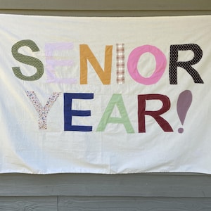 May include: A white fabric banner featuring the words SENIOR YEAR! appliqued in various colorful patterned fabrics. The letters are crafted from different textiles including green polka dot, light purple, mustard yellow, plaid, pink, black polka dot, floral, blue, light green, and red plaid.