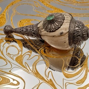 May include: An ornate, antique conch shell with silver-toned metal accents and a central green stone. The shell is cream-colored, and the metalwork features intricate floral designs. The object is reflected on a mirrored surface with gold swirls.