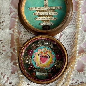 Pode incluir: Uma pequena caixa redonda de madeira com uma tampa com dobradiças. A tampa está aberta e revela um pequeno recipiente redondo de vidro com uma imagem em forma de coração no seu interior. A caixa é decorada com uma cruz e o texto "Zoet Hart van Jesus, maak dat ik U altijd meer en meer beminne. 1300 dag. afl S. Coleta".