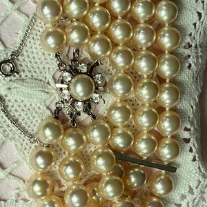 Puede incluir: Una pulsera de perlas vintage con múltiples hilos de perlas de imitación de color crema. La pulsera presenta un cierre decorativo plateado con una perla central rodeada de pedrería transparente. La pulsera está sobre un tapete de encaje blanco.