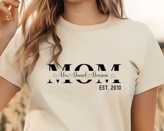 Può includere: Una maglietta bianca con testo nero che dice "MOM" con i nomi "Alex, Daniel, Dominic" e "EST. 2010" sotto.