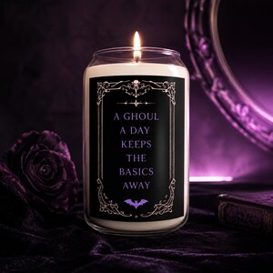 May include: A white wax candle in a clear glass jar with a black label featuring a decorative frame, a small purple bat, and the text A GHOUL A DAY KEEPS THE BASICS AWAY. The candle has a lit wick and is set against a dark purple background.