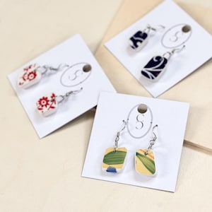 May include: Three pairs of earrings displayed on white cards with the letter "S" printed on them. The earrings are made from ceramic tiles with different patterns. One pair features a red and white floral design, another has a blue and white floral design, and the third pair has a yellow and green floral design.