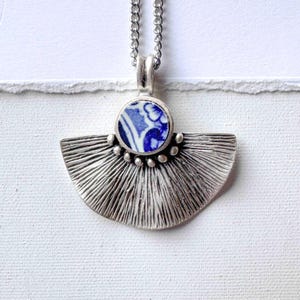 May include: A silver-toned pendant necklace with a fan-shaped design. The pendant features a circular blue and white ceramic piece at the top, with a fan-shaped, textured silver base. The necklace hangs on a delicate silver chain.