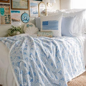 Op de afbeelding: Een bed met wit beddengoed, versierd met kussens en een blauw-wit patroon deken. Een kussen heeft een blauw Amerikaans vlagontwerp. Een houten bordje leest "BEACH". De kamer is ingericht met kunstwerken met kustthema.
