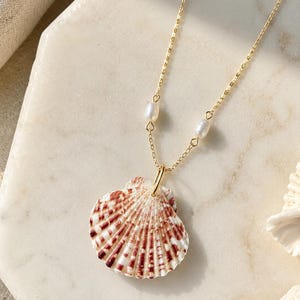 May include: A gold-colored necklace featuring a seashell pendant with red and white markings. The necklace includes two small, white pearls and a delicate chain. The seashell pendant is the focal point, showcasing natural patterns and textures.