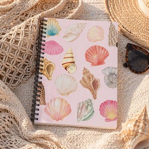 May include: A spiral-bound notebook with a pink cover featuring watercolor illustrations of various seashells in shades of pink, beige, and blue. The notebook rests on a beige knitted blanket, with a straw hat and sunglasses in the background.