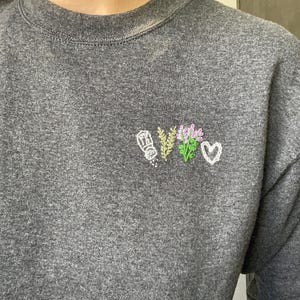 May include: A heather gray sweatshirt with embroidered floral and heart designs. The embroidered elements include a salt shaker, wheat, lavender, and a white heart. The sweatshirt is made of a soft, comfortable material.