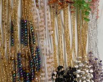 Puede incluir: Una colección de cadenas de cintura con cuentas de varios colores y estilos. Las cadenas presentan diferentes tipos de cuentas, incluyendo oro, cristal y piedras naturales.