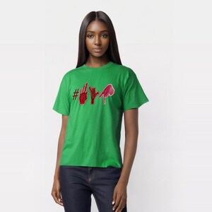 Puede incluir: Una camiseta verde con un gráfico rojo de gestos con las manos que deletrean una palabra. La camiseta está hecha de un material suave y la lleva una persona. El gráfico incluye un símbolo de hashtag.