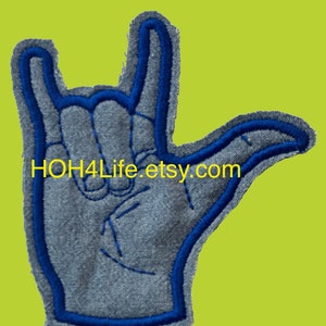 May include: A gray patch in the shape of a hand making the 'I love you' sign, with blue embroidered outlines and details. The text "HOH4Life.etsy.com" is printed in yellow. The patch is set against a lime green background.