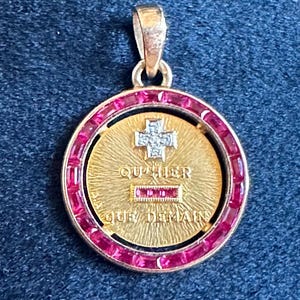 May include: A gold pendant with a red ruby border and a gold cross with a diamond centre. The pendant has the inscription "QU'IL NE FAILLE QU'UN JOUR" in French.