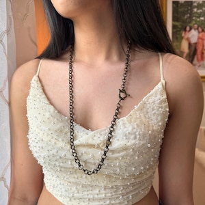 May include: A long, dark metal chain necklace with a toggle clasp. The necklace is worn over a cream-colored, beaded top. The background includes a window with a patterned curtain and a framed photograph.