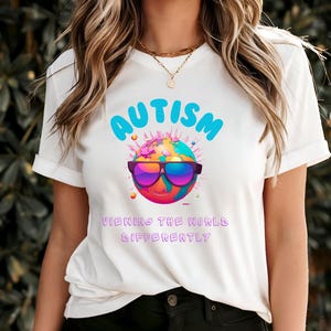 May include: White t-shirt with a blue and pink graphic of a smiling earth wearing sunglasses. The text "AUTISM VIEWING THE WORLD DIFFERENTLY" is printed on the shirt.
