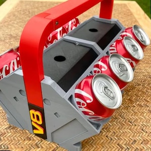 May include: A gray and red 3D printed can holder with a handle. The holder has six slots for cans and the text "VB" printed on the side.