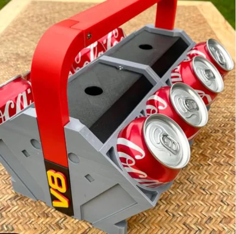 May include: A gray and red 3D printed can holder with a handle. The holder has six slots for cans and the text "VB" printed on the side.