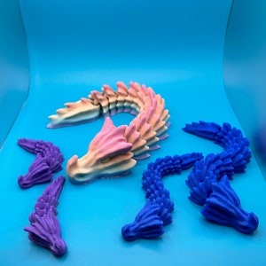 May include: A collection of 3D-printed dragon figures in various colors. The central dragon is a gradient of pink, yellow, and blue. Two blue dragons and two purple dragon heads are also present, all on a blue background.