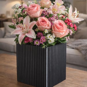 May include: A floral display of pink roses, lilies, and other flowers in a black, vertically-lined square vase. The flowers are in full bloom, creating a vibrant display. The vase sits on a wooden table.