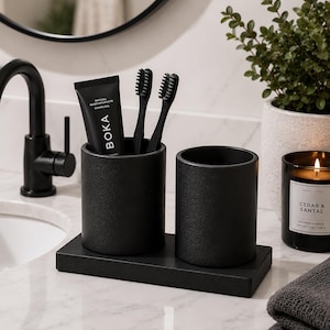 May include: A black bathroom set comprising a toothbrush holder, two toothbrushes, and a toothpaste tube. The toothpaste tube is labelled "BOKA". The set sits on a marble countertop, with a candle and plant visible in the background.