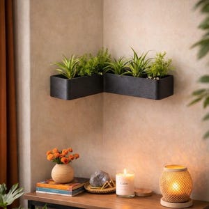 Op de afbeelding: Een zwarte, L-vormige wandplanter gevuld met groene planten. Daaronder staat een houten tafel met een vaas met oranje bloemen, boeken, een kaars met het opschrift "Calm" en een geweven lamp. De scène speelt zich af tegen een neutraal getinte muur.