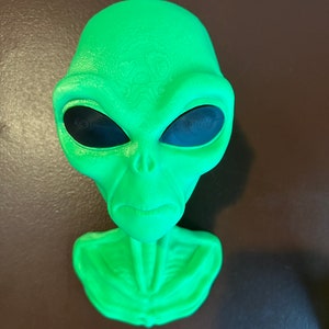 Puede incluir: Una figura de cabeza de extraterrestre verde con ojos negros. El extraterrestre tiene una cara texturizada y un cuello y pecho detallados.
