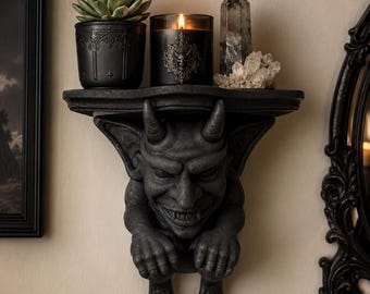 May include: A dark gray wall-mounted shelf shaped like a gargoyle with horns and a wide grin. The shelf holds a black succulent planter, a black candle with a metallic emblem, a tall dark crystal point, and a cluster of clear quartz crystals.