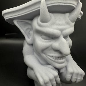 May include: A white, detailed gargoyle sculpture with a pointed hat and a mischievous expression. The gargoyle is crouching with its hands resting on its knees.