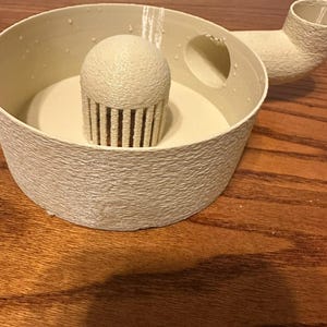 K&ouml;nnte beinhalten: Ein cremefarbenes, strukturiertes, 3D-gedrucktes Objekt mit rundem Sockel, einem zentralen cupcake-f&ouml;rmigen Element und einem gebogenen Ausguss. Das Objekt hat eine raue, por&ouml;se Oberfl&auml;che und scheint ein K&uuml;chen- oder Dekorationsartikel zu sein.