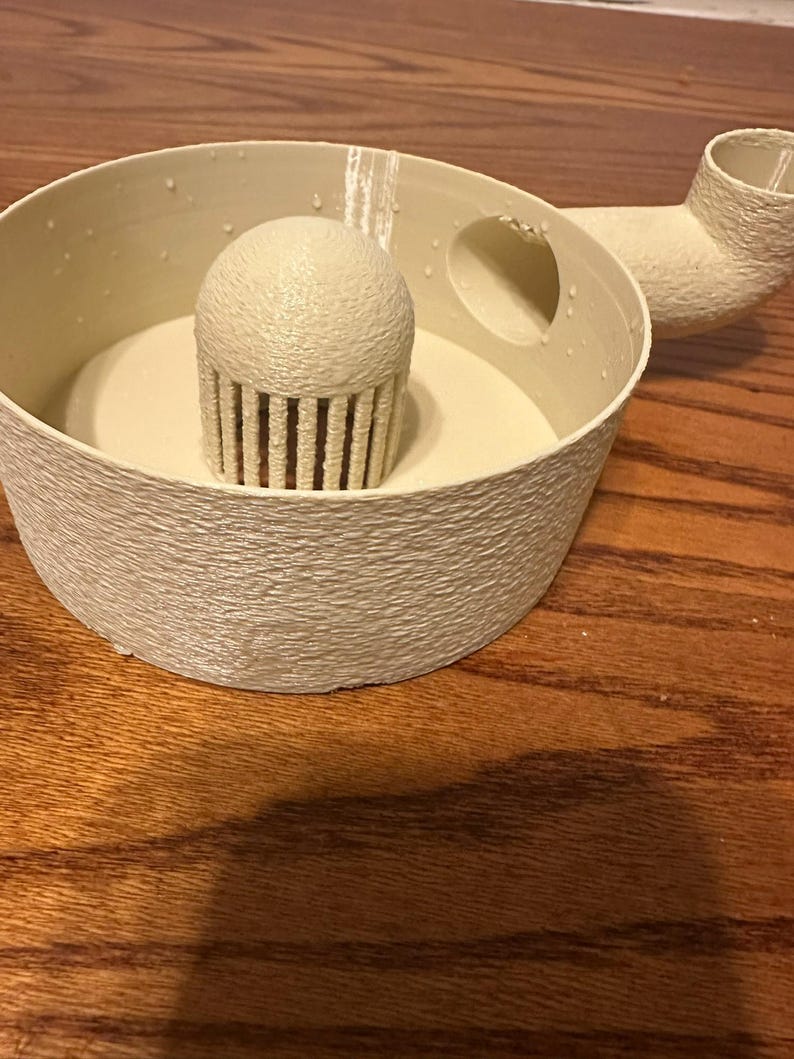 K&ouml;nnte beinhalten: Ein cremefarbenes, strukturiertes, 3D-gedrucktes Objekt mit rundem Sockel, einem zentralen cupcake-f&ouml;rmigen Element und einem gebogenen Ausguss. Das Objekt hat eine raue, por&ouml;se Oberfl&auml;che und scheint ein K&uuml;chen- oder Dekorationsartikel zu sein.
