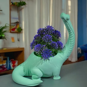 Puede incluir: Un macetero de dinosaurio de color verde claro con un arreglo de flores moradas en su interior. El macetero está sobre una superficie gris.