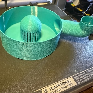 K&ouml;nnte beinhalten: Ein t&uuml;rkisblaues 3D-gedrucktes Objekt mit kreisf&ouml;rmiger Basis, einem zentralen zylindrischen Aufbau mit vertikalen Linien und einem gebogenen Rohr. Das Objekt aus strukturiertem Kunststoff befindet sich auf einer dunklen Oberfl&auml;che.