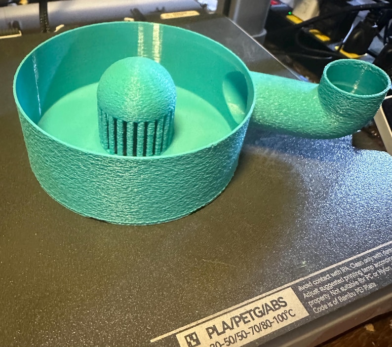K&ouml;nnte beinhalten: Ein t&uuml;rkisblaues 3D-gedrucktes Objekt mit kreisf&ouml;rmiger Basis, einem zentralen zylindrischen Aufbau mit vertikalen Linien und einem gebogenen Rohr. Das Objekt aus strukturiertem Kunststoff befindet sich auf einer dunklen Oberfl&auml;che.