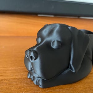 May include: A black 3D printed dog planter. The dog is lying down with its head turned to the side. The planter is made of a smooth, matt black plastic.