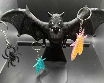 Op de afbeelding: Een zwarte vleermuisvormige displaystandaard met drie hangende sleutelhangers. De linker sleutelhanger is een zwarte spin, de middelste is een blauwgroen insect en de rechter is een roze en geel insect, elk met een metalen ring.