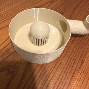 K&ouml;nnte beinhalten: Ein cremefarbenes, 3D-gedrucktes Objekt mit kreisf&ouml;rmiger Basis, einer zentralen, abgerundeten Struktur mit vertikalen Lamellen und einem angeschlossenen Rohr. Das Objekt liegt auf einer Holzoberfl&auml;che. Das Design scheint ein Filter oder Sieb zu sein.
